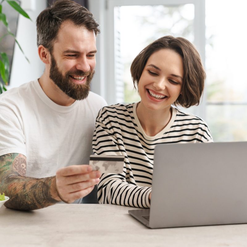 Portrait of a cheerful couple shopping online with laptop computer while sitting at the table in a kitchen at home, man holding credit card